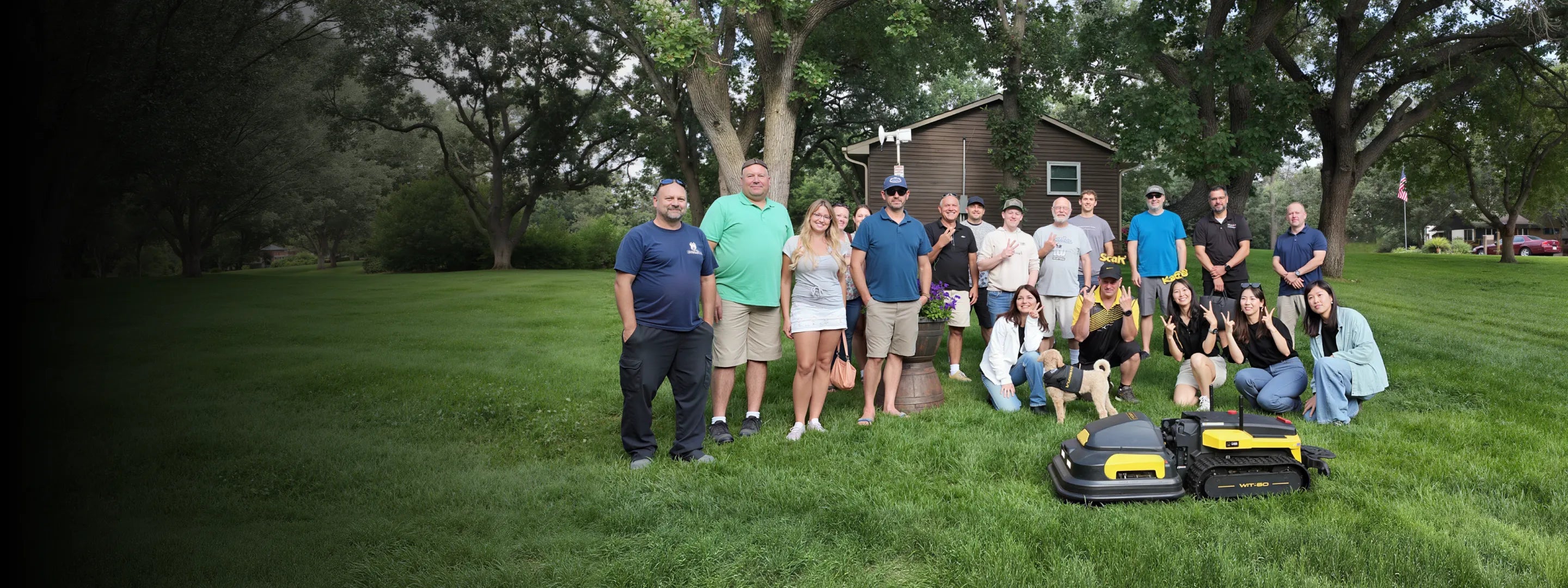 Group of people posing on a grassy lawn with two robotic lawn mowers and a small dog in front, trees and a shed in the background.