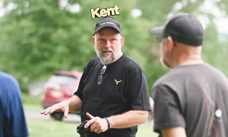 Man with gray beard and black cap labeled "Kent" gestures while speaking outdoors, wearing black shirt with sunglasses and gold logo.