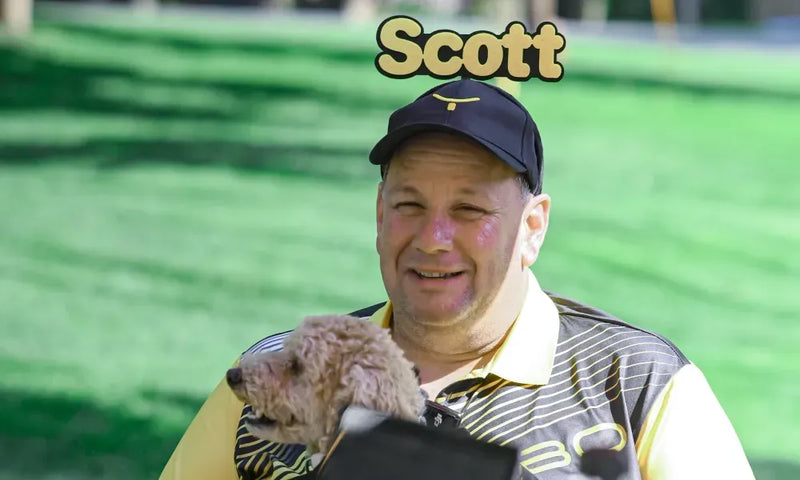 Man named Scott smiles while holding a small fluffy dog in a carrier, wearing a black cap and striped polo shirt, outdoors on green grass.