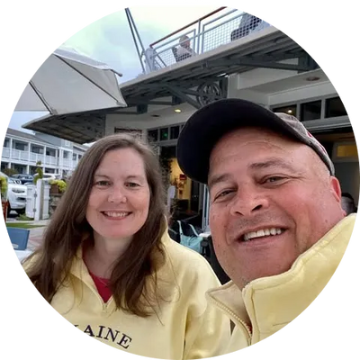 Smiling couple in yellow jackets at outdoor waterfront cafe, man wearing cap, woman with long brown hair, umbrellas and building in background.