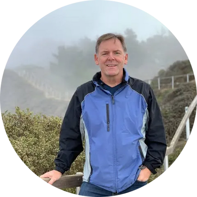 Man in blue and black jacket smiles outdoors on foggy hillside, hand on railing, trees and fence in background.