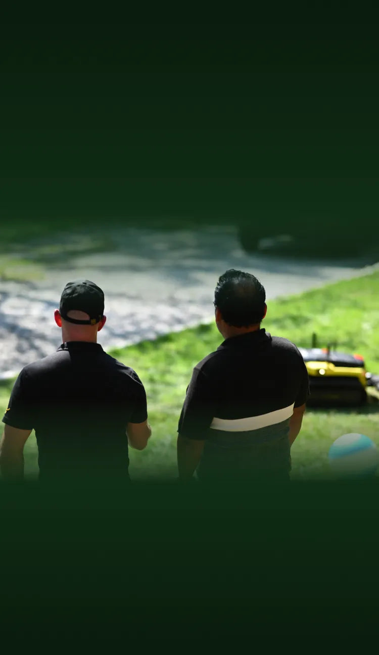 Two men in black shirts stand back-to-back on grass, facing a gravel path and yellow equipment under bright sunlight.