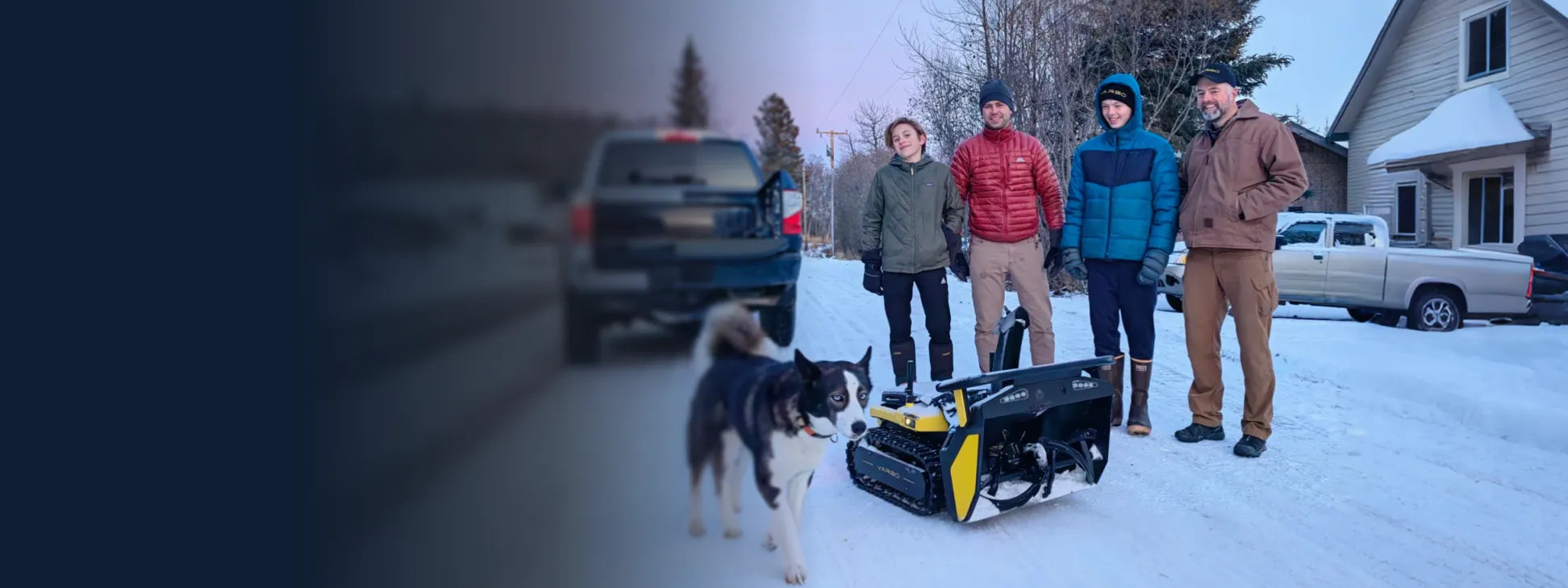 Four people in winter gear stand in snow with a black and white dog and a yellow-and-black tracked robot, trucks and house in background.