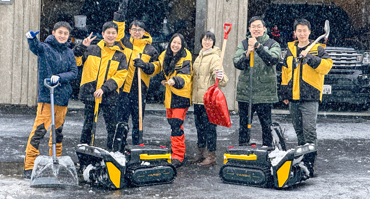 Seven people in winter gear pose with shovels and YARBO snow-clearing robots in falling snow, smiling and gesturing.