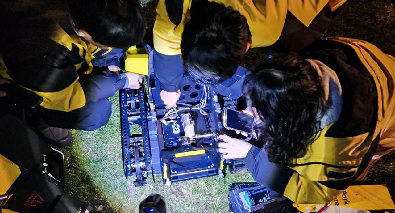 Three people in yellow jackets work on a blue robotic vehicle on grass at night, using tools and a phone.