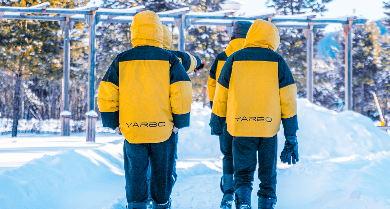 Four children in yellow and black YARBO winter jackets walk through snow under a wooden pergola, backs to camera.