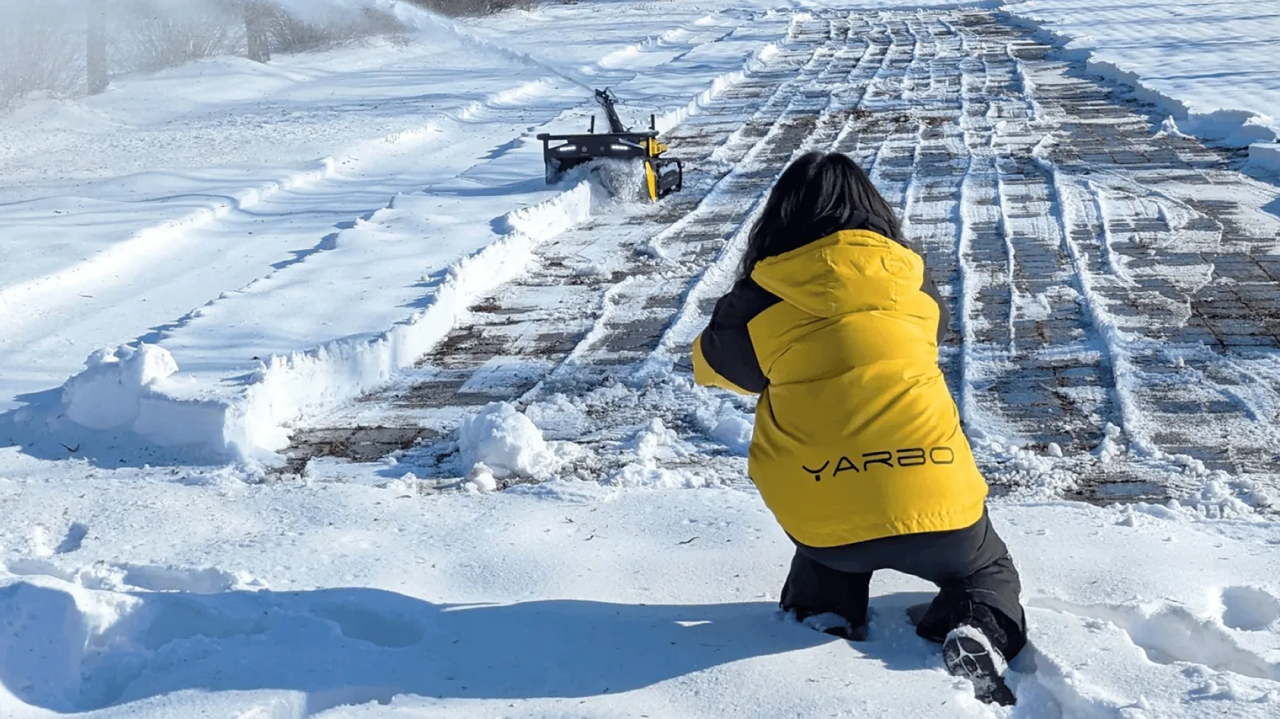 Person in yellow YARBO jacket kneels in snow, watching snowblower clear brick path. Bright sunlight, winter scene.