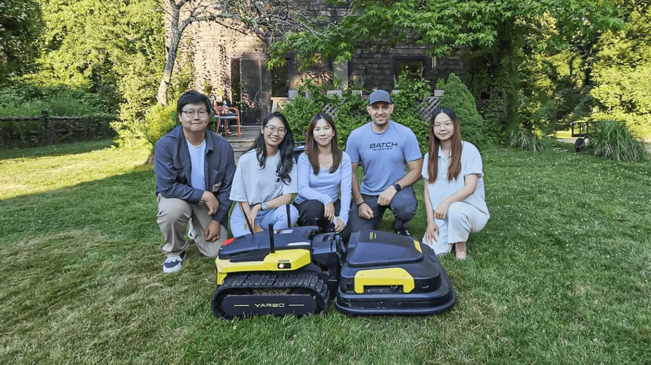 Five people crouch on a grassy lawn behind two black and yellow YARBO robotic lawn mowers, with a stone house and greenery in the background.