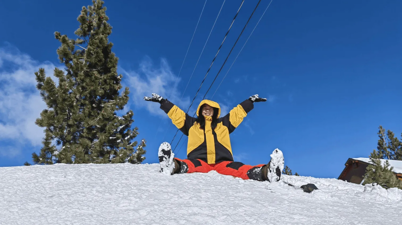 Person in yellow jacket and red pants sits in snow with arms outstretched, smiling under blue sky with evergreen tree and ski lift cables.
