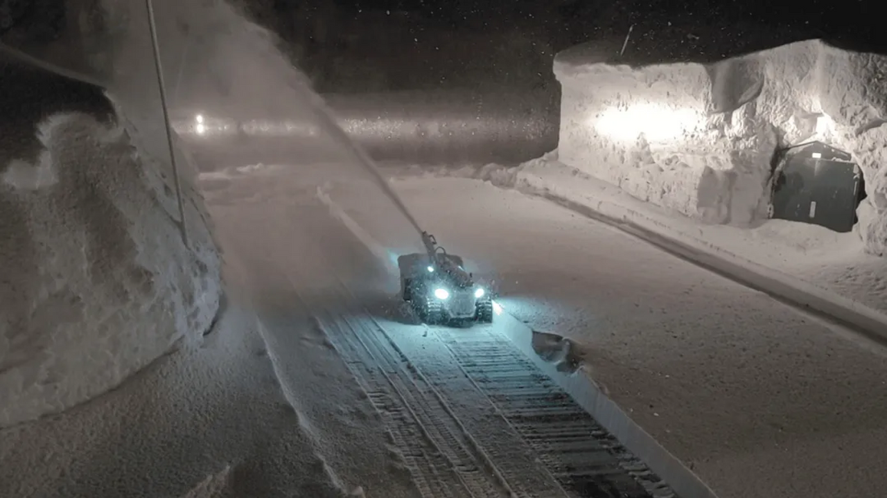Snowplow clears driveway at night, headlights illuminating snow spray, high snowbanks on sides.
