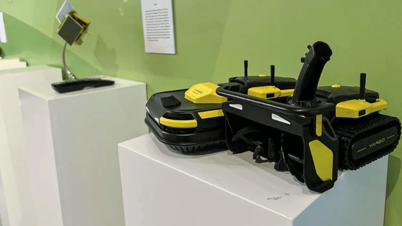 Black and yellow YARD robotic lawn mowers on white display pedestals against a green wall.