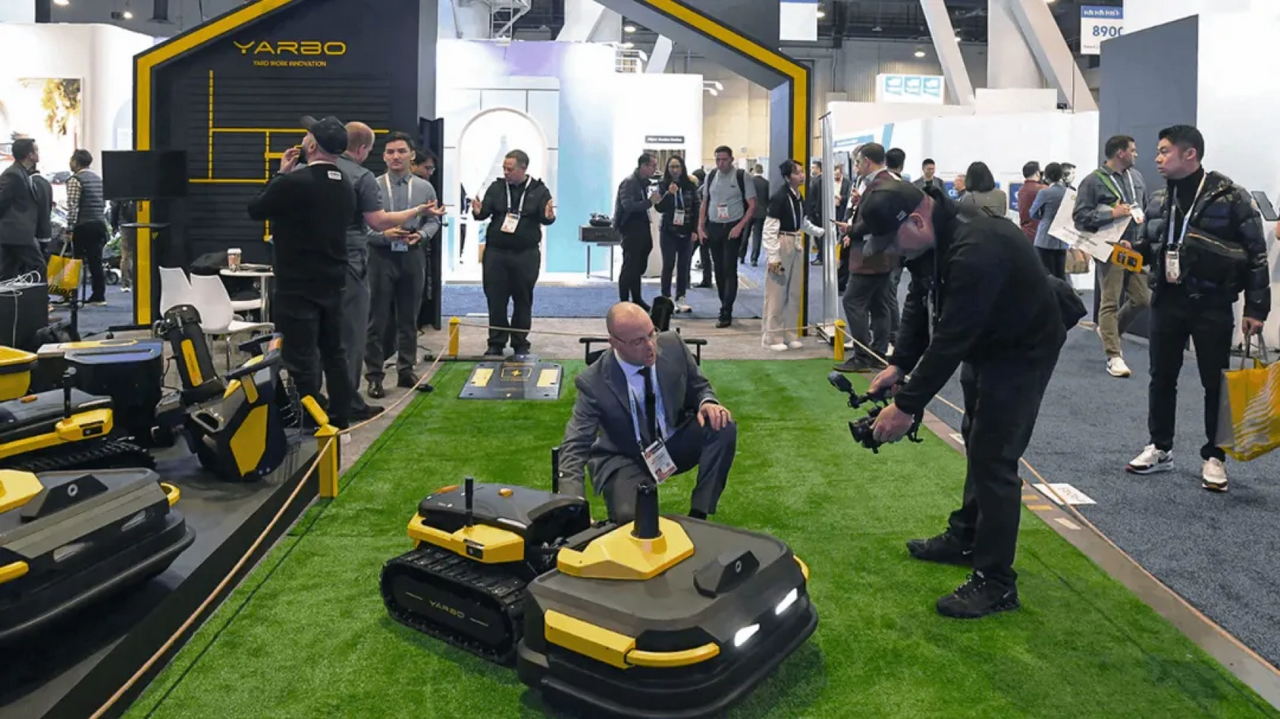 Yellow and black YARBO robotic mowers on green turf at trade show, with attendees observing and photographing.