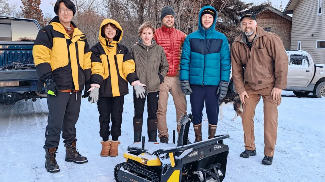 Six people in winter jackets stand in snow beside a yellow and black tracked robot, with trucks and a house in the background.