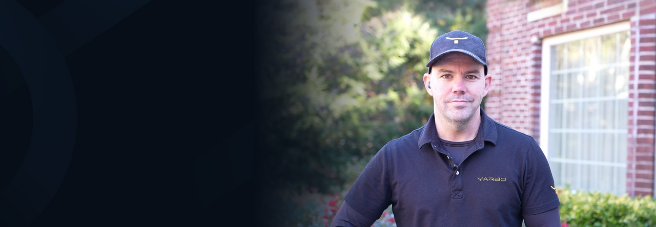 Man in black polo shirt with "YARBO" logo and matching cap stands outdoors near brick building with window, greenery in background.