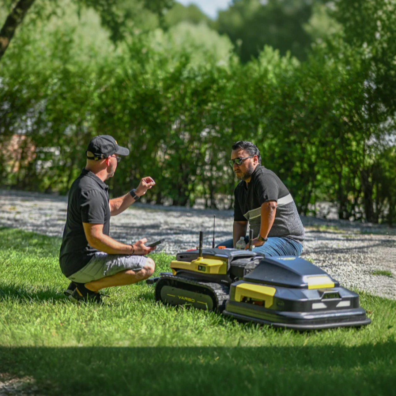 Two men crouch on grass beside yellow and black robotic lawn mowers, discussing features in a sunny outdoor setting.