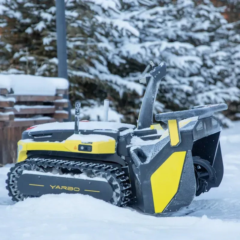 Yellow and black YARBO robotic lawn mower on snow-covered ground, tracks visible, snow-dusted evergreens in background.