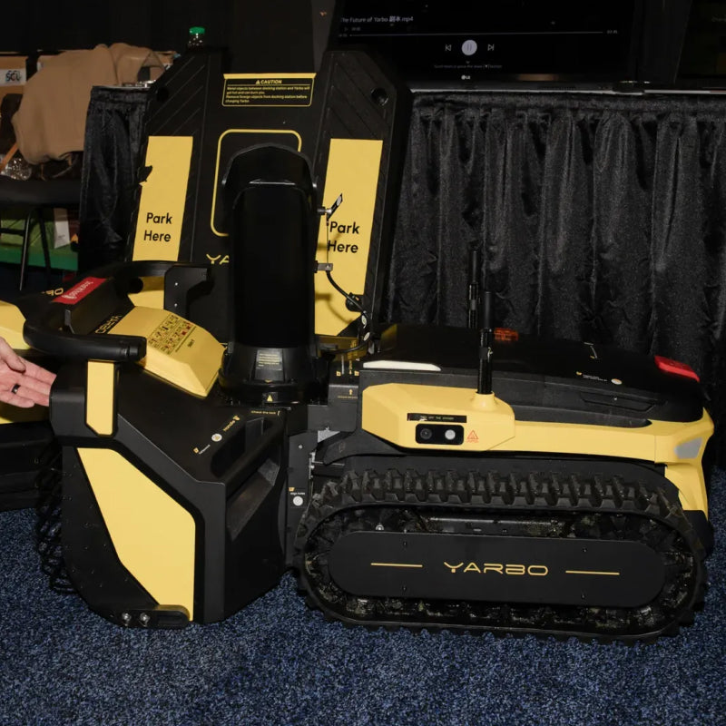 Yellow and black YARBO robotic lawn mower with tank treads, parked at charging station with "Park Here" labels, on blue carpet.