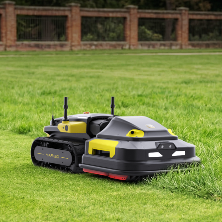 Black and yellow robotic lawn mower on green grass, featuring tracks and antennas, with brick fence in background.