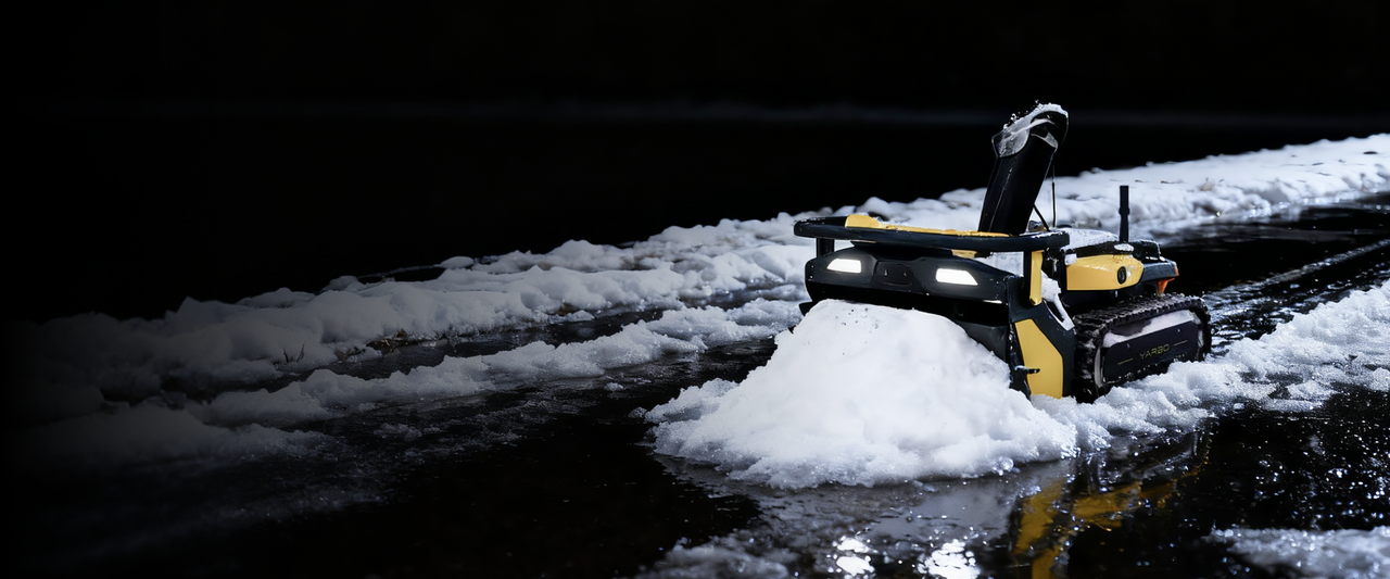 Yellow and black YARBO robotic snow blower clearing snow at night, illuminated by headlights on wet ground.