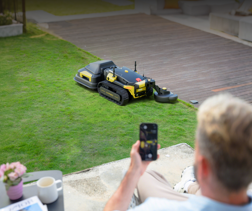 Man relaxes on patio, using smartphone to control black and yellow robotic lawn mower on green grass.
