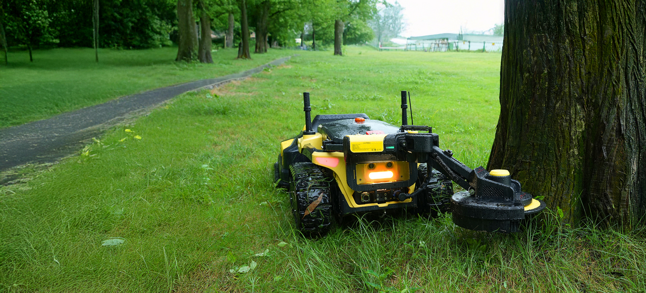 Yellow and black tracked robot with illuminated front light and mechanical arm, positioned on grass beside a tree trunk in a park.