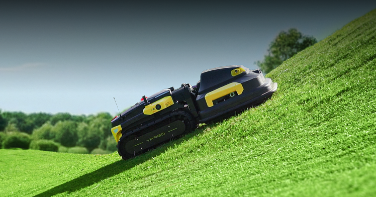 Black and yellow VARBO robotic lawn mower on a grassy slope under a clear sky.