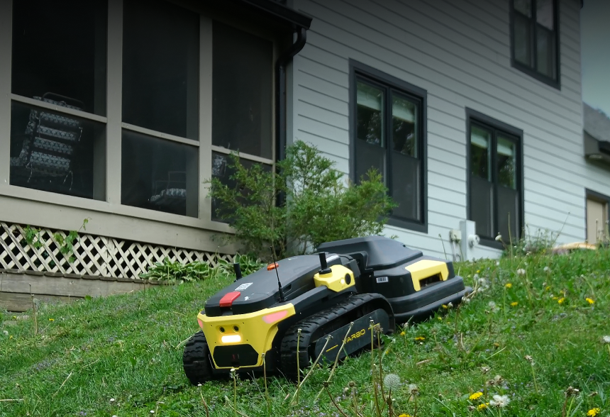 Yellow and black robotic lawn mowers on grass near a house with gray siding and dark windows.