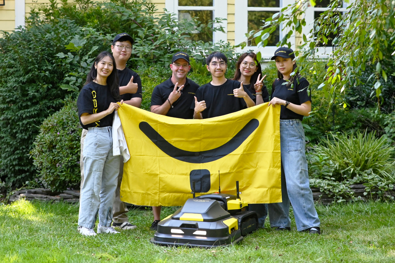 Six people in black shirts and caps hold a yellow smiley-face banner outdoors, standing behind two black and yellow robotic lawn mowers on green grass.