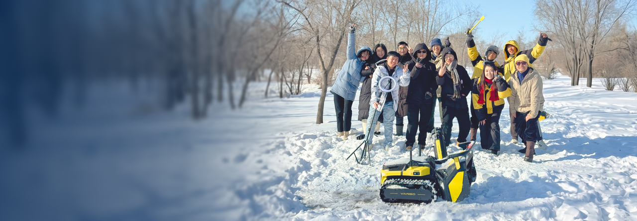 Group of people in winter coats pose joyfully in snowy park with yellow and black tracked robot in foreground.