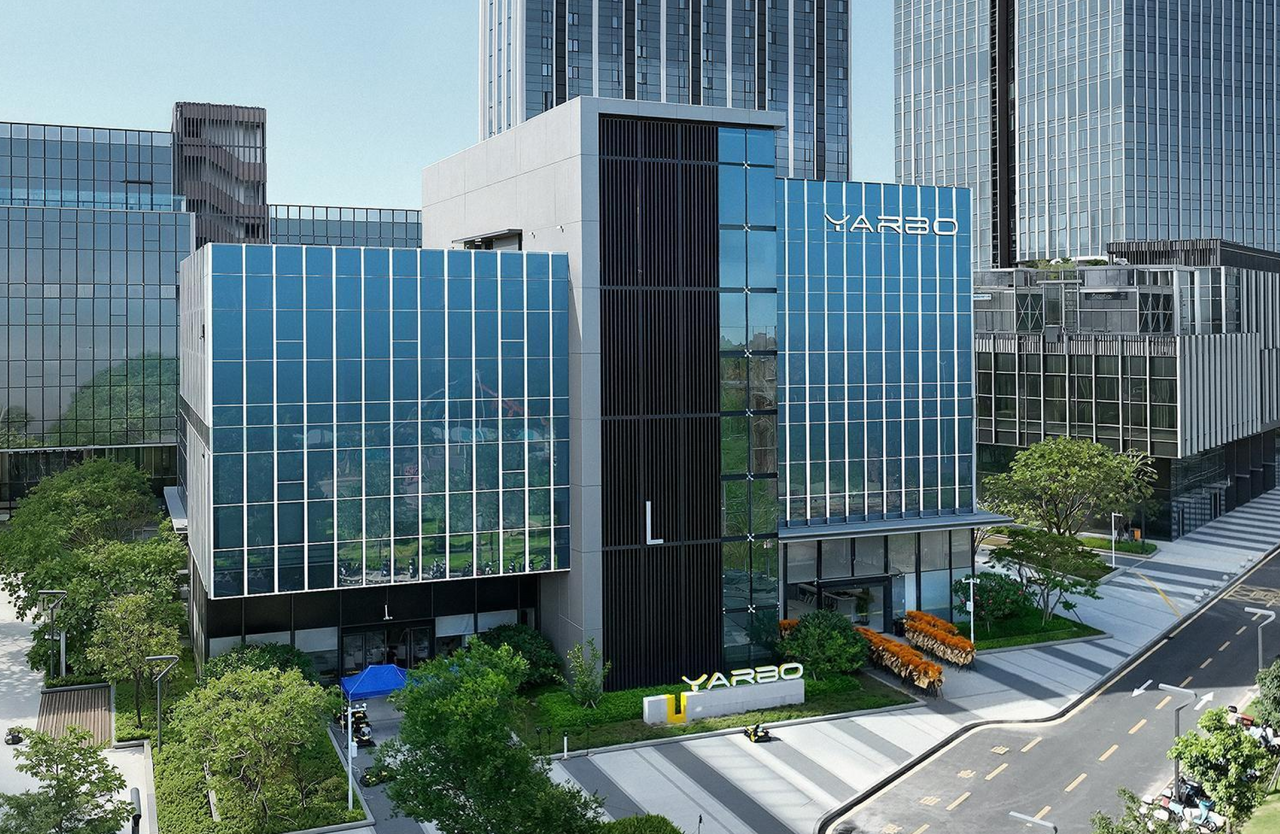 Modern glass YARBO building with blue facade, surrounded by greenery and tall skyscrapers under clear sky.