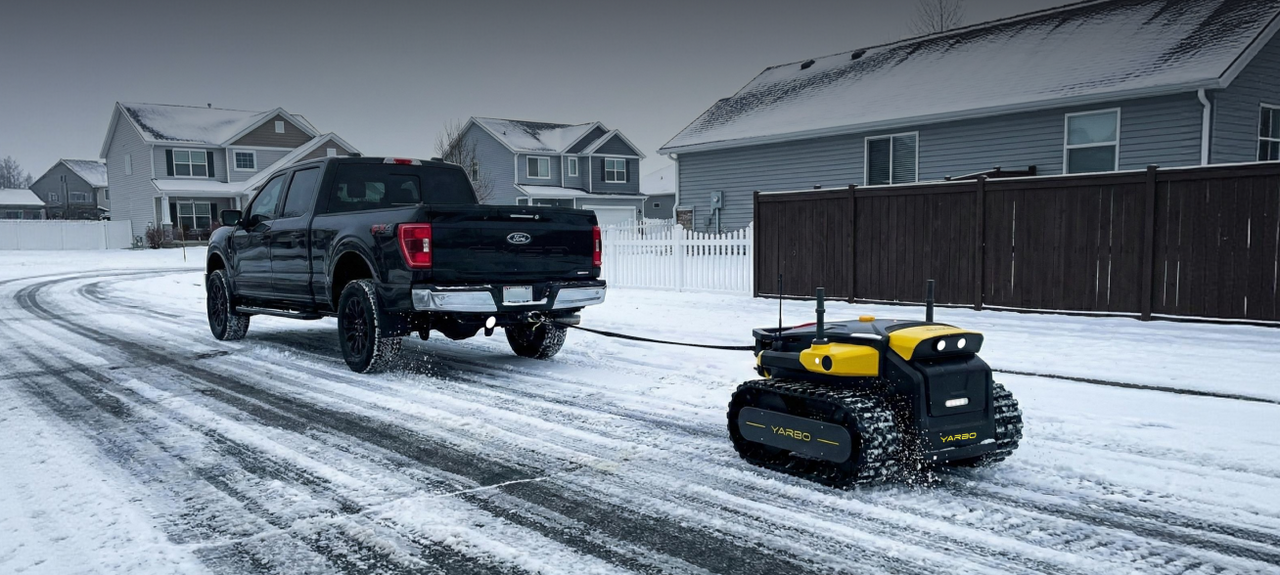 Black Ford truck towing yellow and black YARBO snowplow robot on snowy suburban driveway.