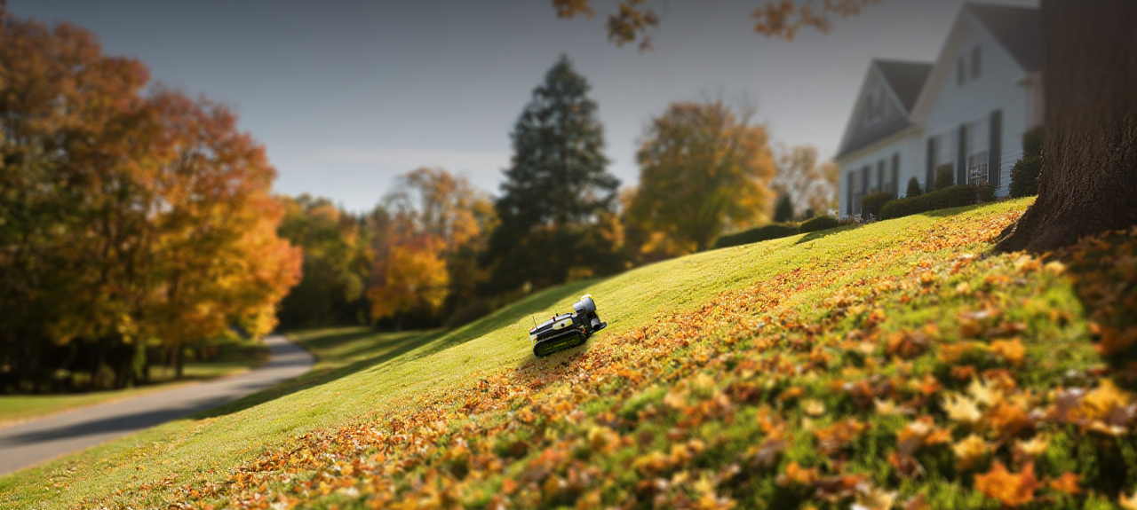 Robotic lawn mower on green grass with autumn leaves, near white house and colorful trees.