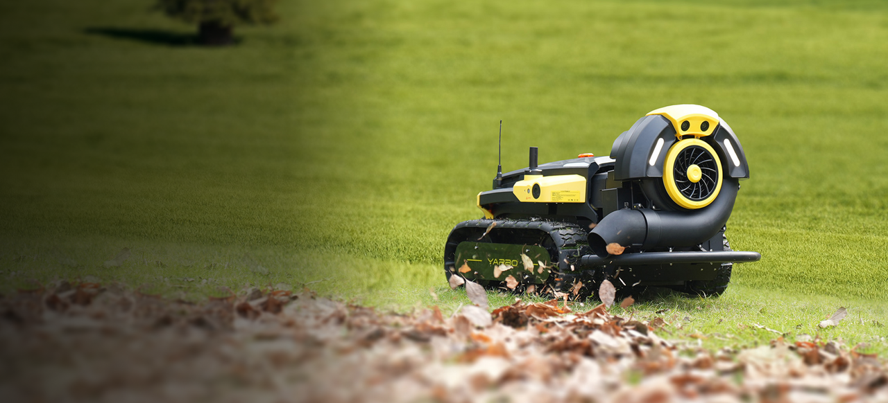 Yellow and black robotic lawn mower with tracks blowing leaves on green grass.