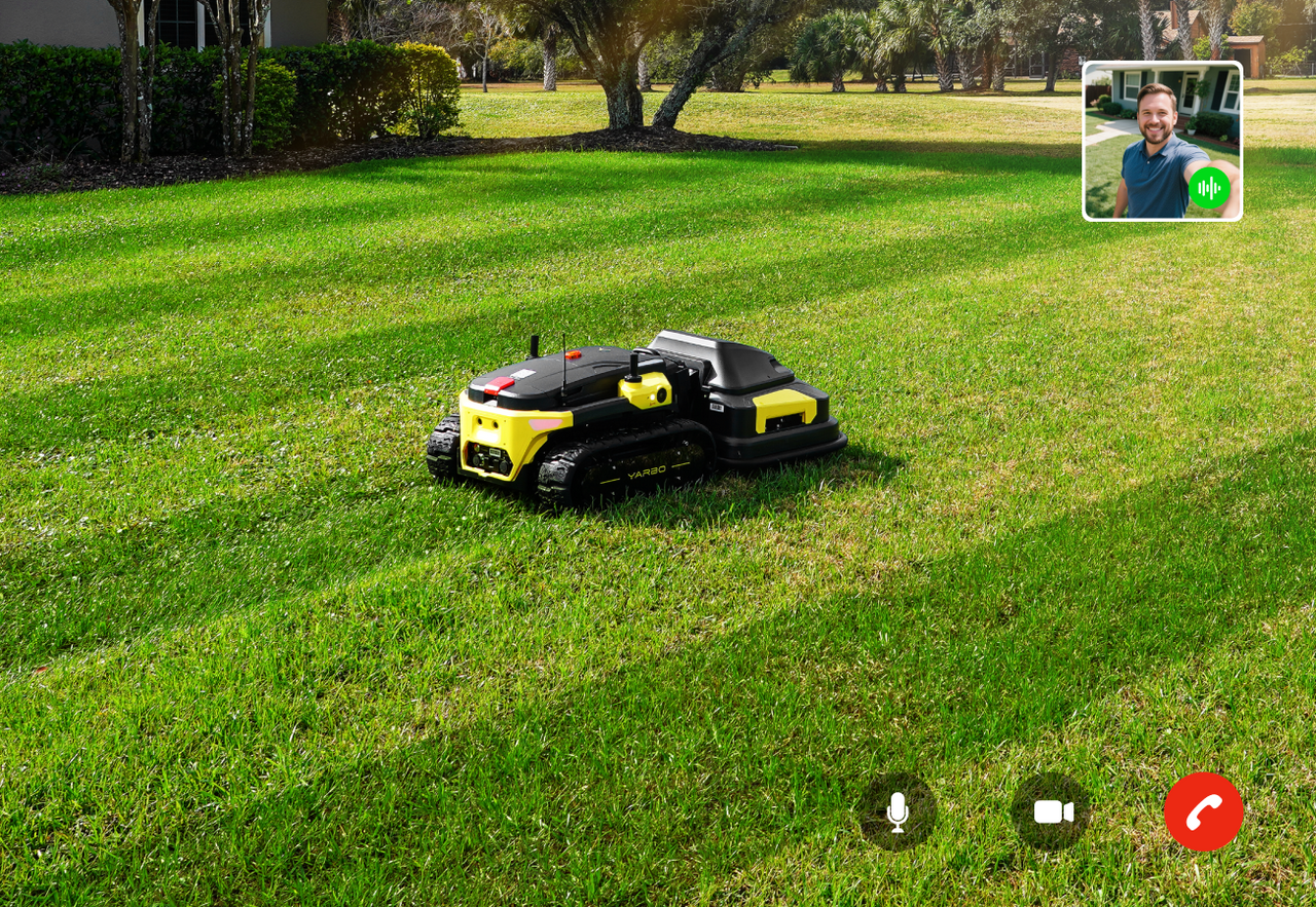 Black and yellow robotic lawn mower on green grass with video call interface overlay showing a smiling man in top right corner.