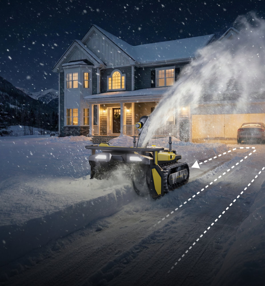 Yellow and black robotic snow blower clears driveway at night in front of a warmly lit house with snow falling.