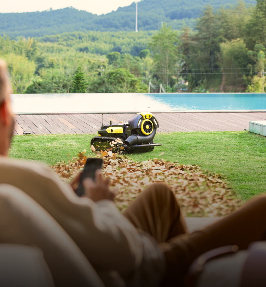 Man relaxes on lounge chair, watching black and yellow robotic lawn mower collect leaves on green grass near pool and forested hills.