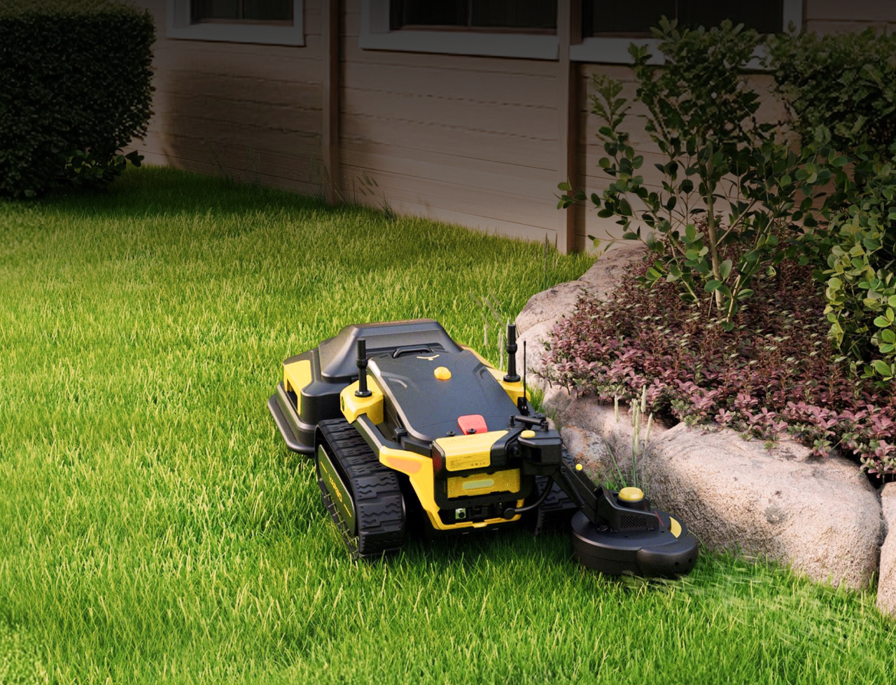 Yellow and black robotic lawn mower on green grass near rocks and shrubs, house in background.