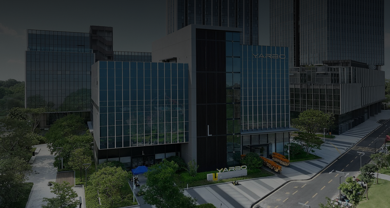 Modern glass YARBO building with green landscaping, surrounded by tall urban structures under overcast sky.