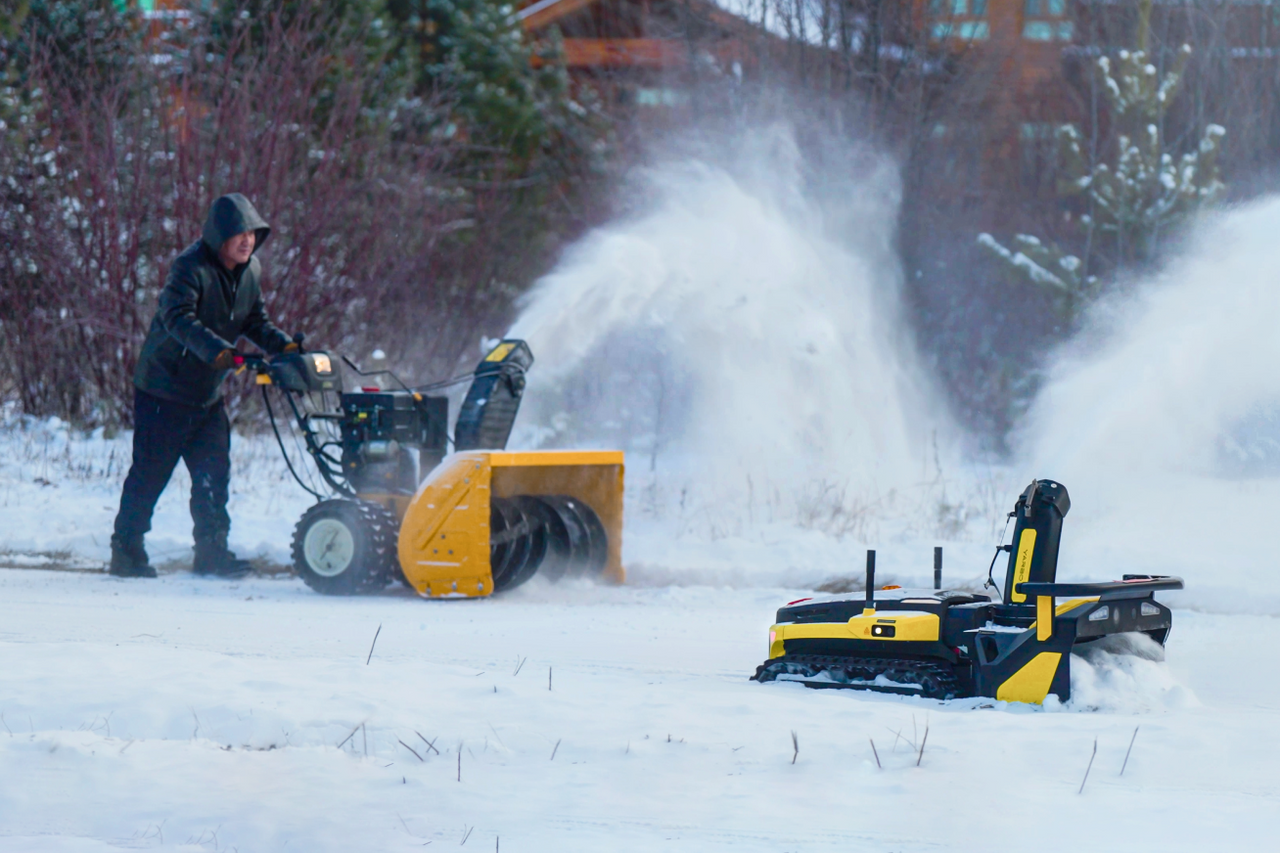 Man operates yellow snowblower, clearing snow in winter yard; robotic snowblower nearby.