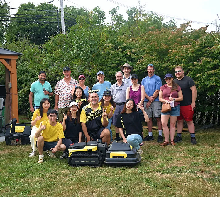 Group of people posing outdoors with two yellow and black robotic lawn mowers in foreground, green trees and fence in background.