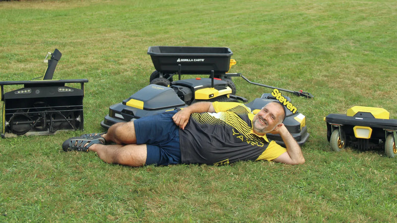 Man named Steven lies on grass surrounded by yellow and black lawn equipment, smiling at camera.
