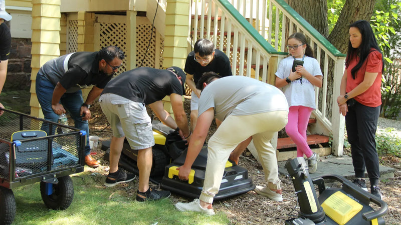Four men assembling yellow and black lawnmowers on grass near a house, with two women observing nearby.