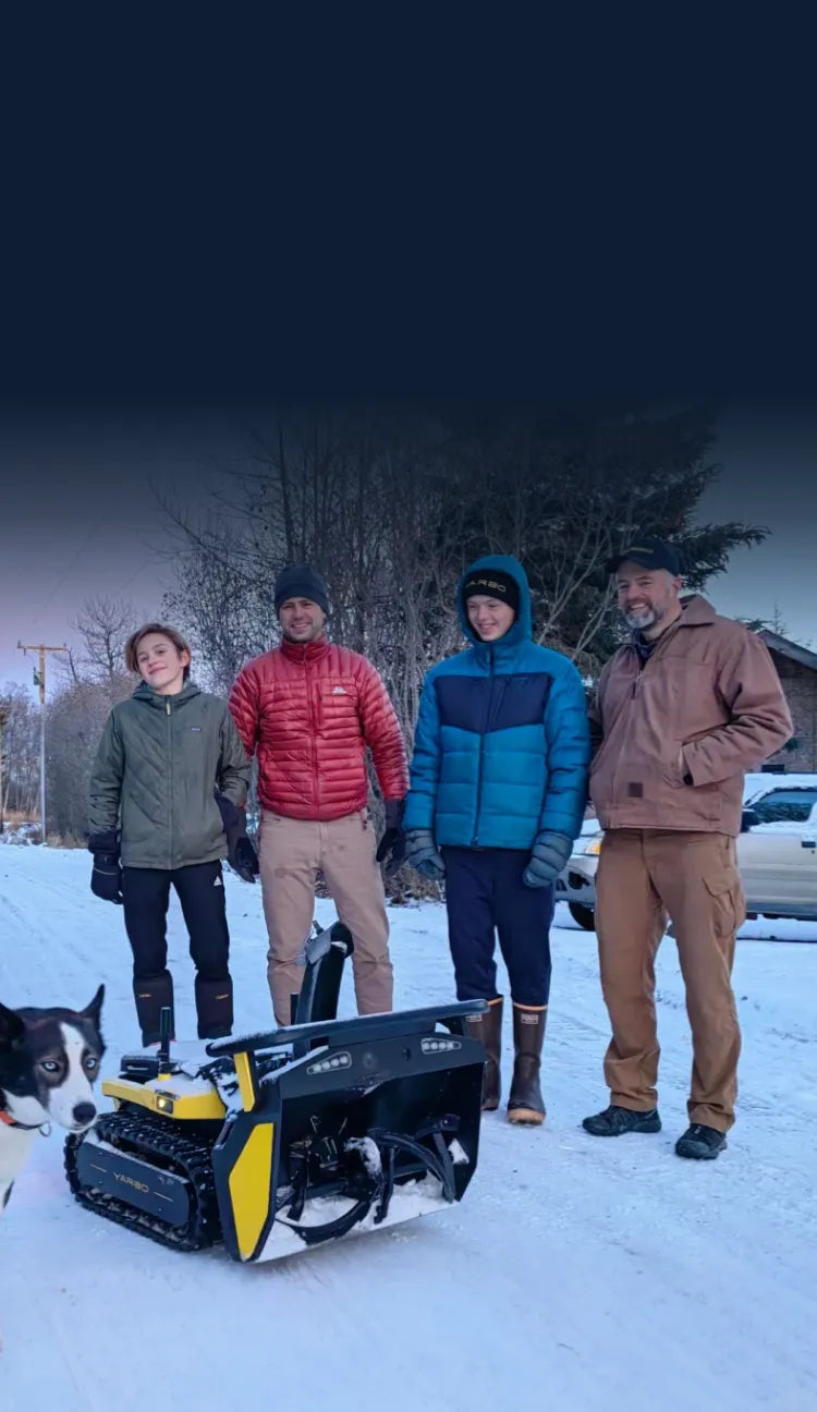 Four people in winter jackets stand in snow with a yellow and black snowblower and a black-and-white dog in foreground.