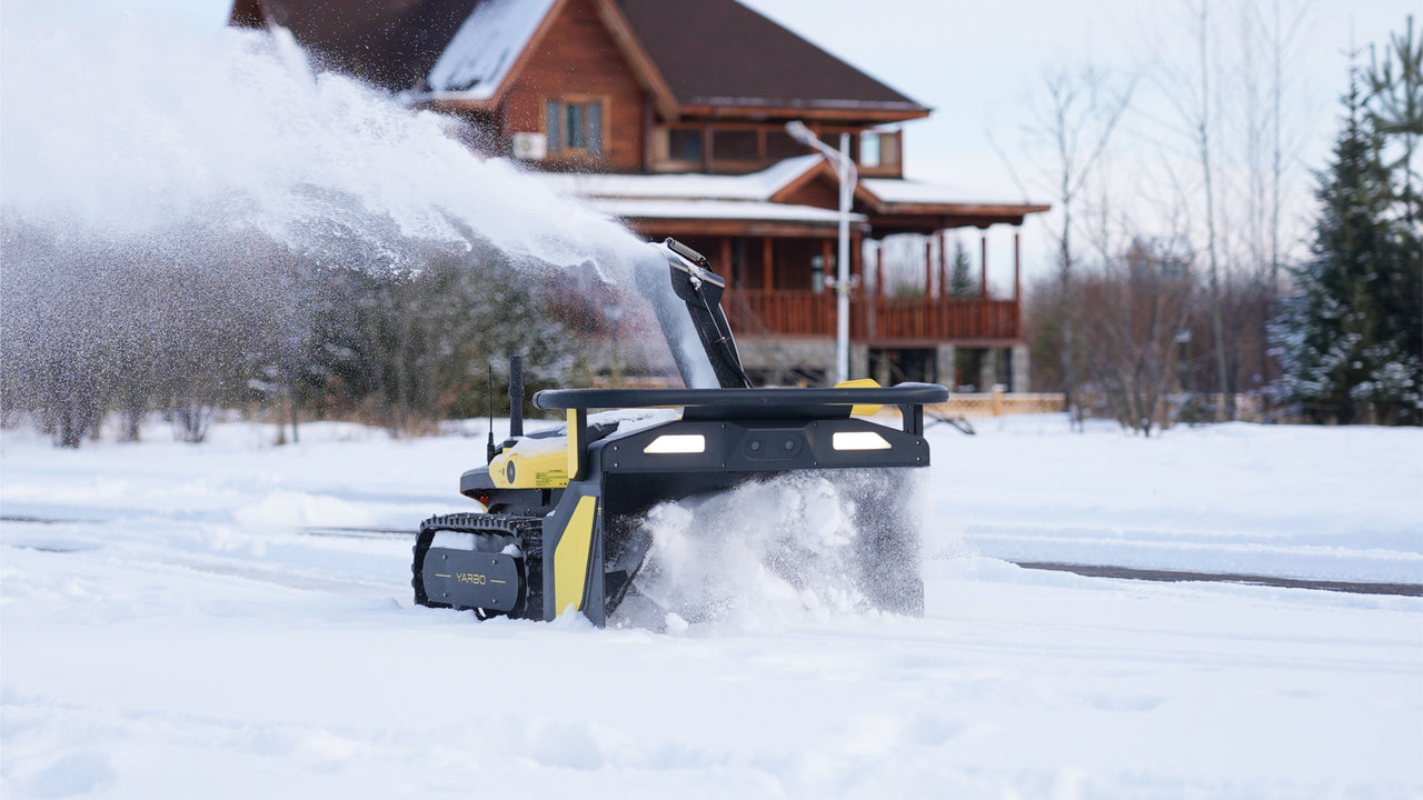Yellow and black snow blower module by YARBO clearing snow in a snowy yard, side view with tracks and illuminated front panel.