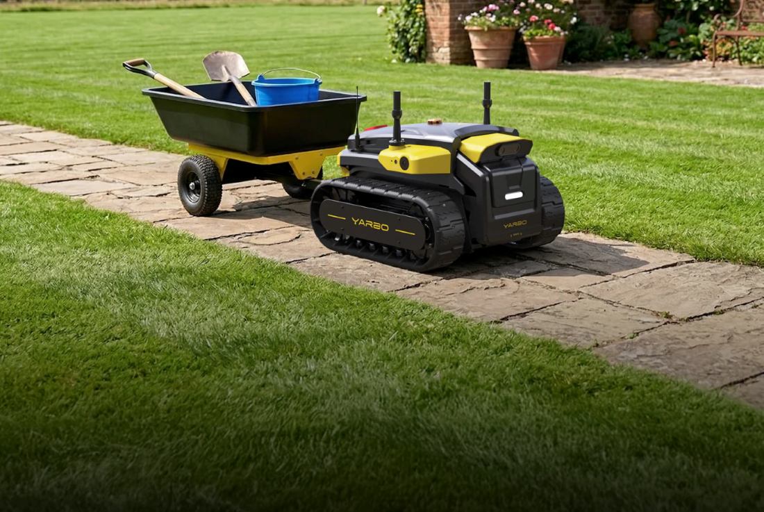Black and yellow YARBO robotic lawn mower on stone path beside green lawn, near wheelbarrow with blue bucket and shovel.