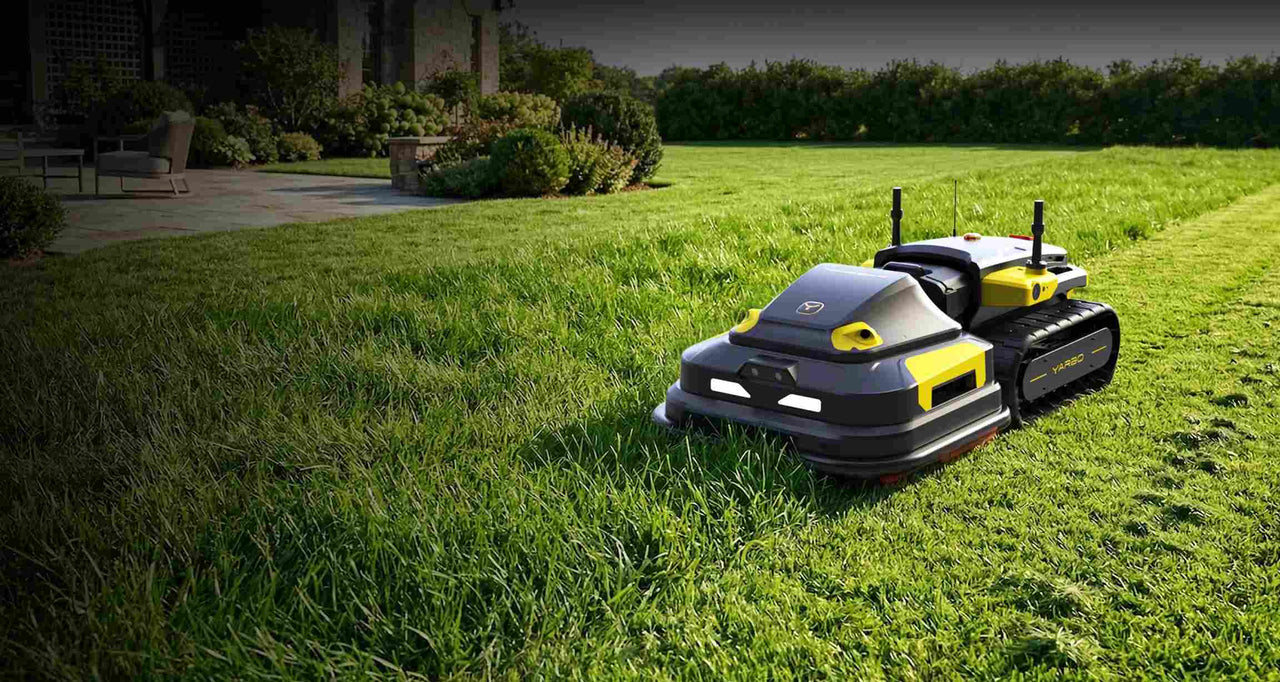Black and yellow robotic lawn mower on lush green grass near a patio with shrubs and a house in the background.
