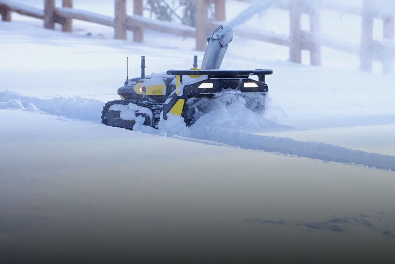 Yellow and black snow-clearing robot with tracks plows snow, spraying white powder, wooden fence in background.