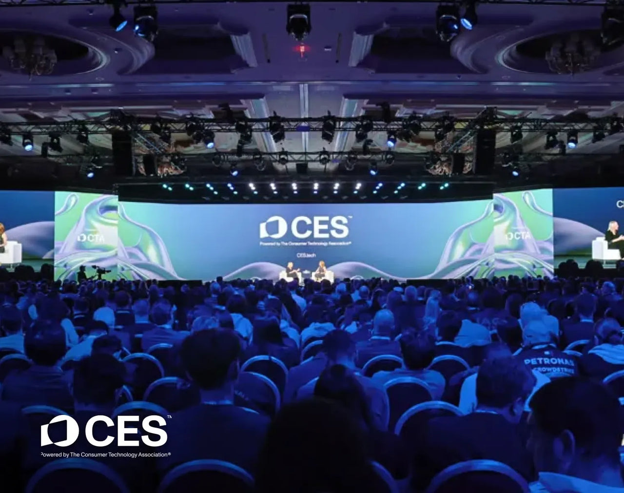 Large audience at CES conference, seated facing stage with CES logo, blue lighting, and speakers on stage.