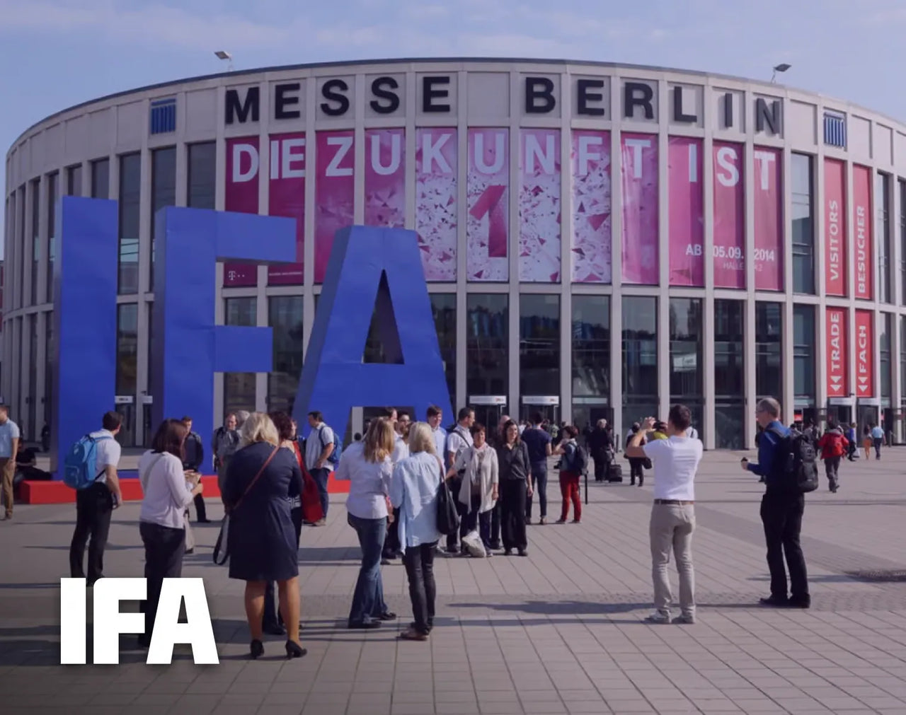 Large blue IFA letters in front of Messe Berlin building, people gathering outside, pink banners reading "Die Zukunft ist" and "IFA 2014".