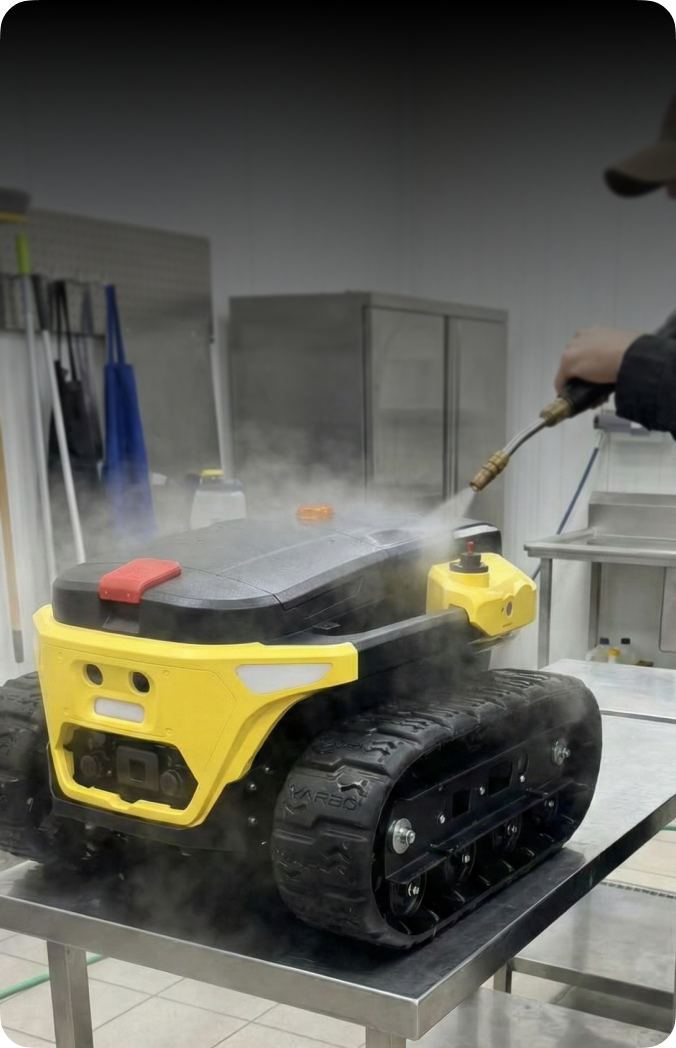 Yellow and black tracked robot being cleaned with steam on metal table in workshop.